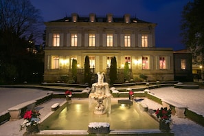 Un imposant bâtiment doté d’une fontaine centrale, entouré de parterres de jardin recouverts de neige et de haies taillées.