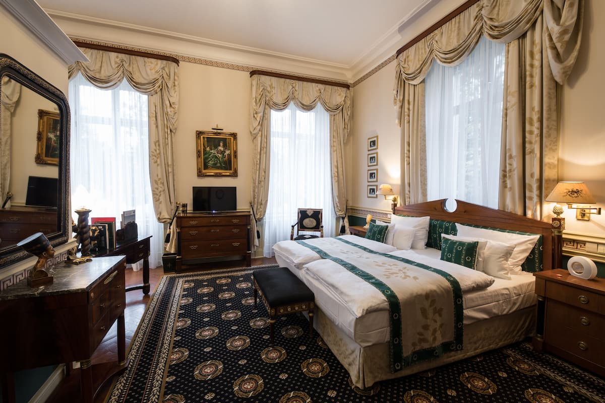 A traditional hotel room with a large bed, a desk, a chair, a dresser, a mirror, and a window with curtains.