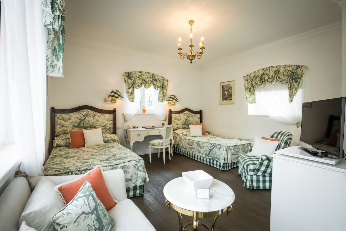 A hotel room with two beds, a desk, a chair, a sofa, a TV, and a chandelier.