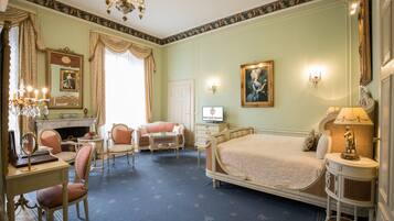 A classic room with a bed, two chairs, a desk, a mirror, a fireplace, and wall sconces.