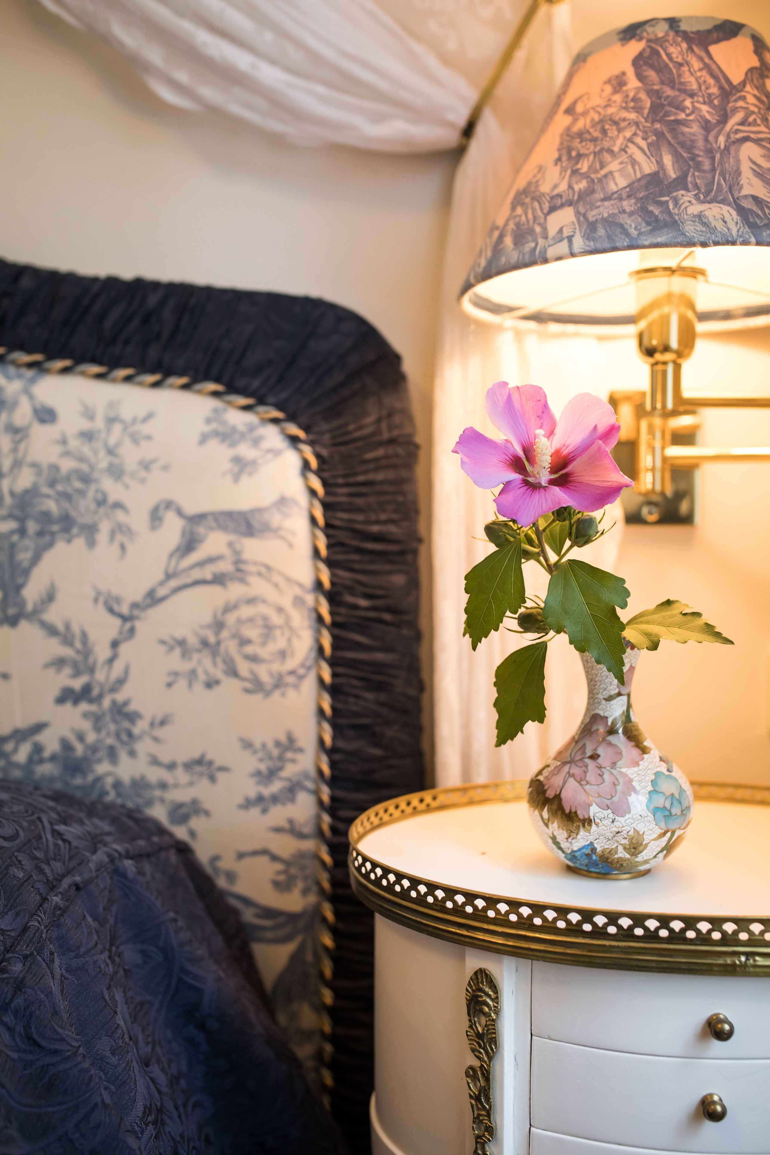 Una cama con colcha azul estampada, una mesita de noche blanca con un florero de flores rosas, y una lámpara con pantalla azul estampada.