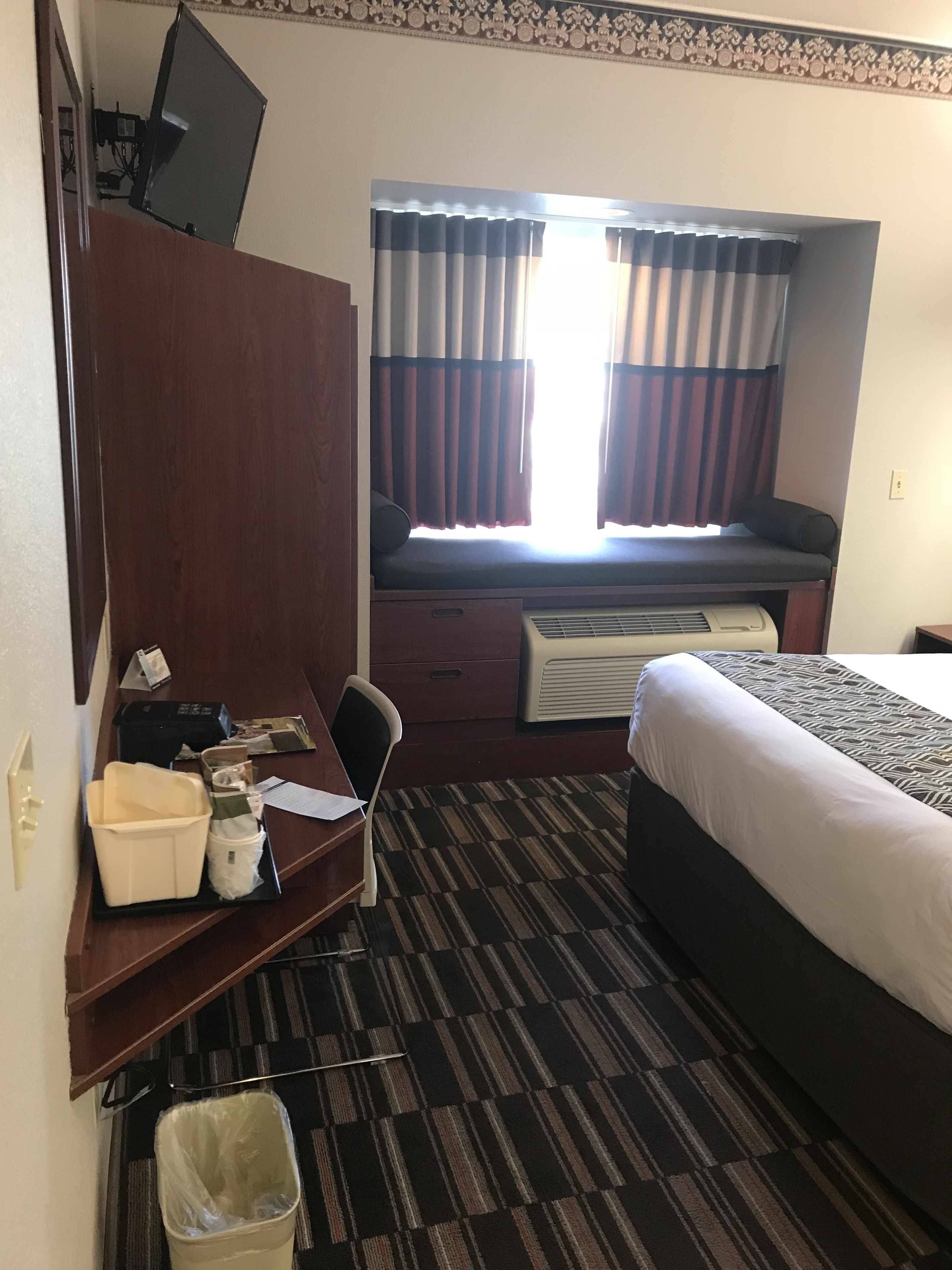 A hotel room with a bed, a desk with a computer, a chair, a waste bin, and a window with curtains.