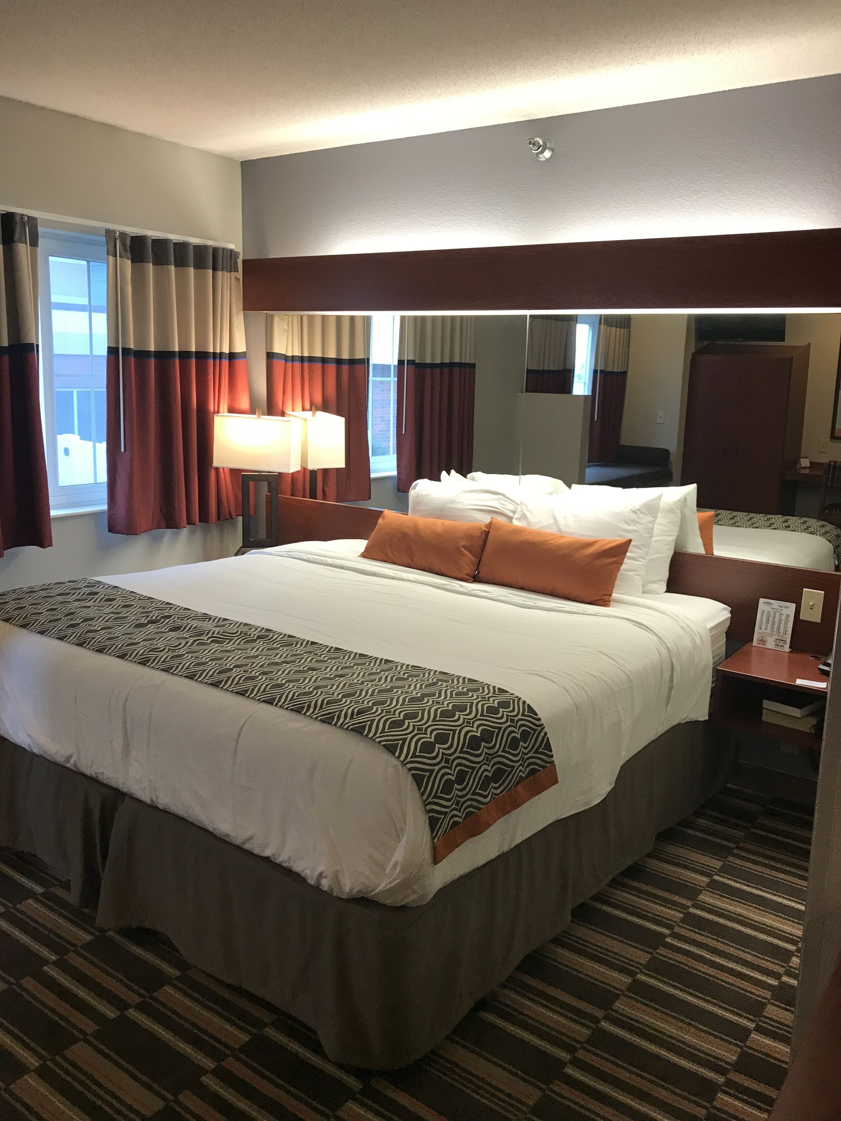 A hotel room with a large bed, a bedside table, a lamp, a mirror, and a window with curtains.