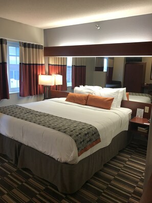Habitación de hotel con una cama grande, una mesita de noche, una lámpara, un espejo y una ventana con cortinas.