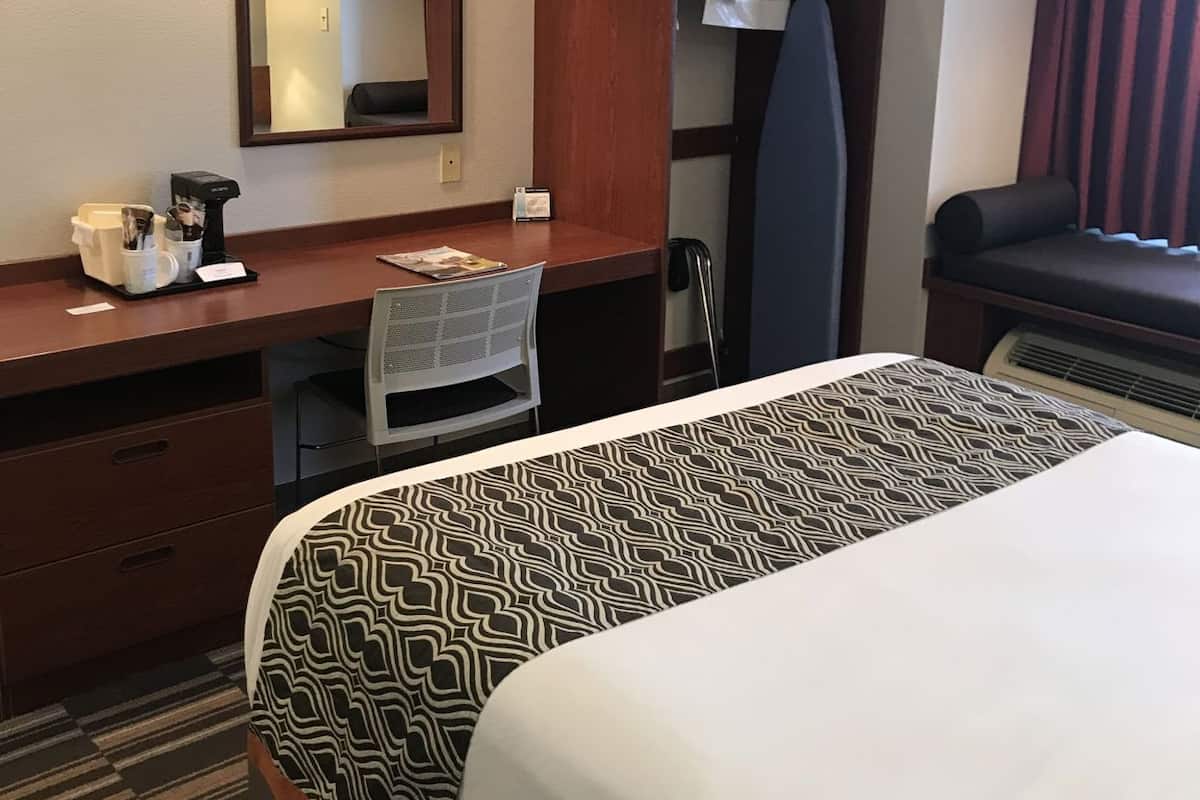 Une chambre d’hôtel comprenant un lit, un bureau avec une chaise, un miroir, une télévision et une fenêtre avec des rideaux.