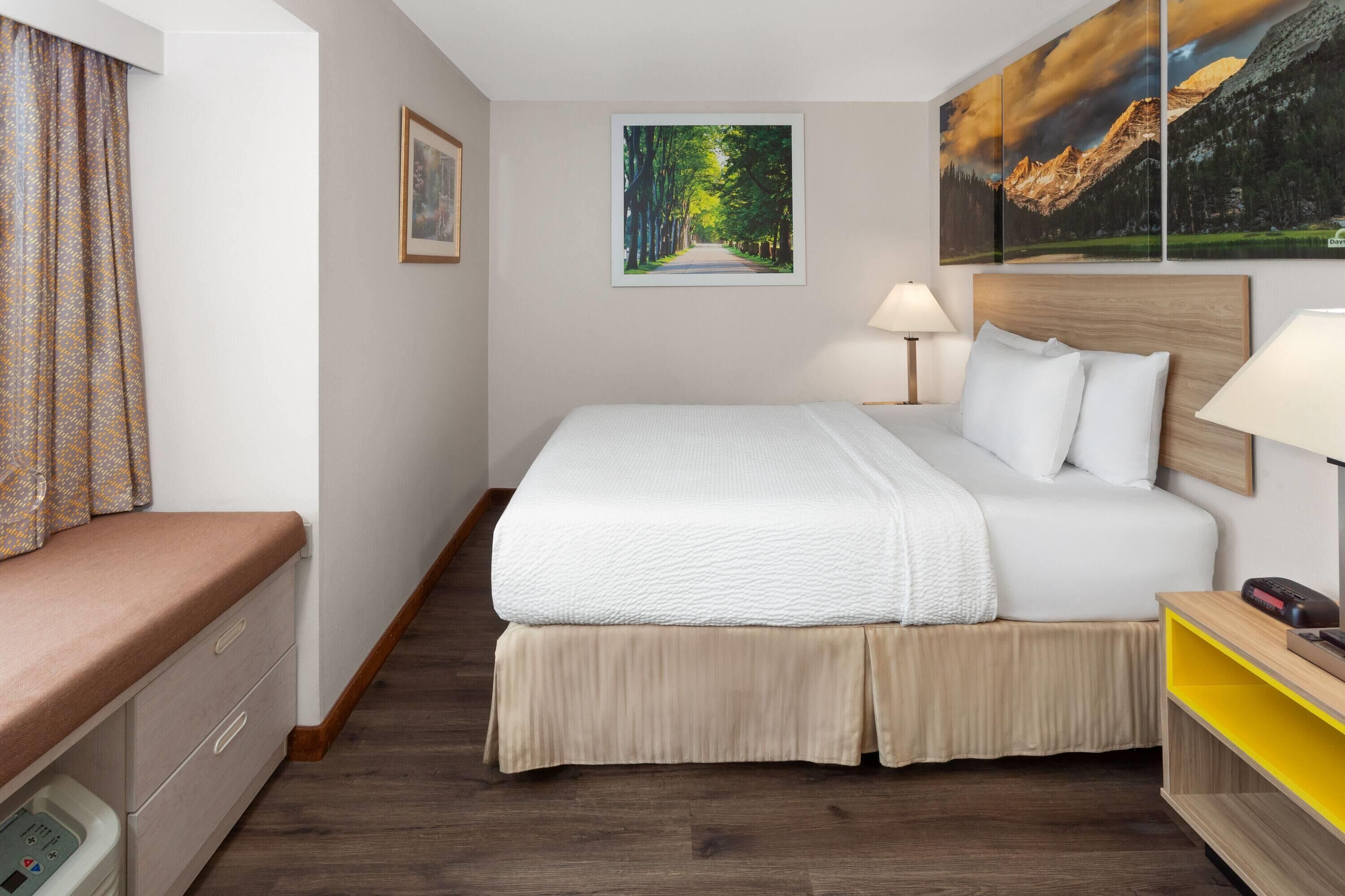 Habitación de hotel con una cama grande, lámparas de noche, cabecera de madera, mesita de noche pequeña, una silla, una ventana con cortinas y un cuadro en la pared.