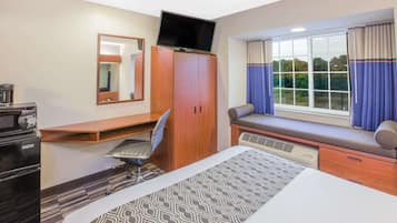 Hotellihuone, jossa on sänky, työpöytä tuolilla, televisiotaulutaso, ikkuna verhoilla ja näkymä puita kohti.