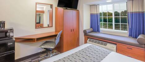 A hotel room with a bed, a desk with a chair, a television on a cabinet, a window with curtains, and a view of trees outside.