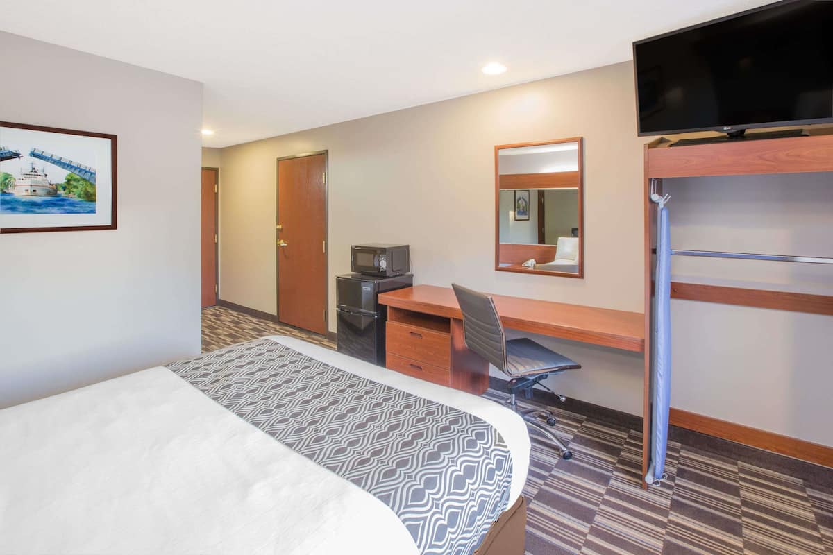A hotel room with a bed, desk, chair, television, and a painting of a bridge.