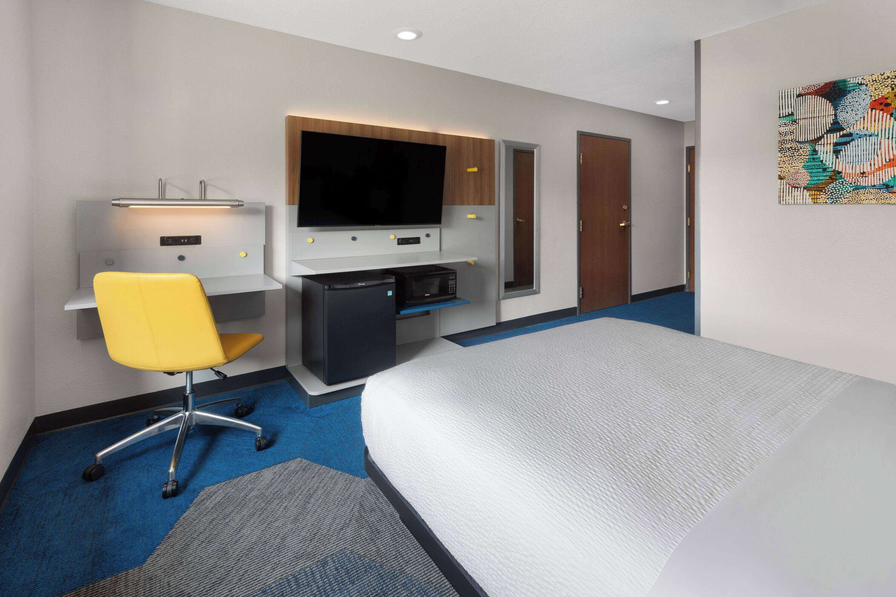 Une chambre d’hôtel avec un lit, un bureau doté d’un téléviseur à écran plat, un four à micro-ondes et une chaise jaune.
