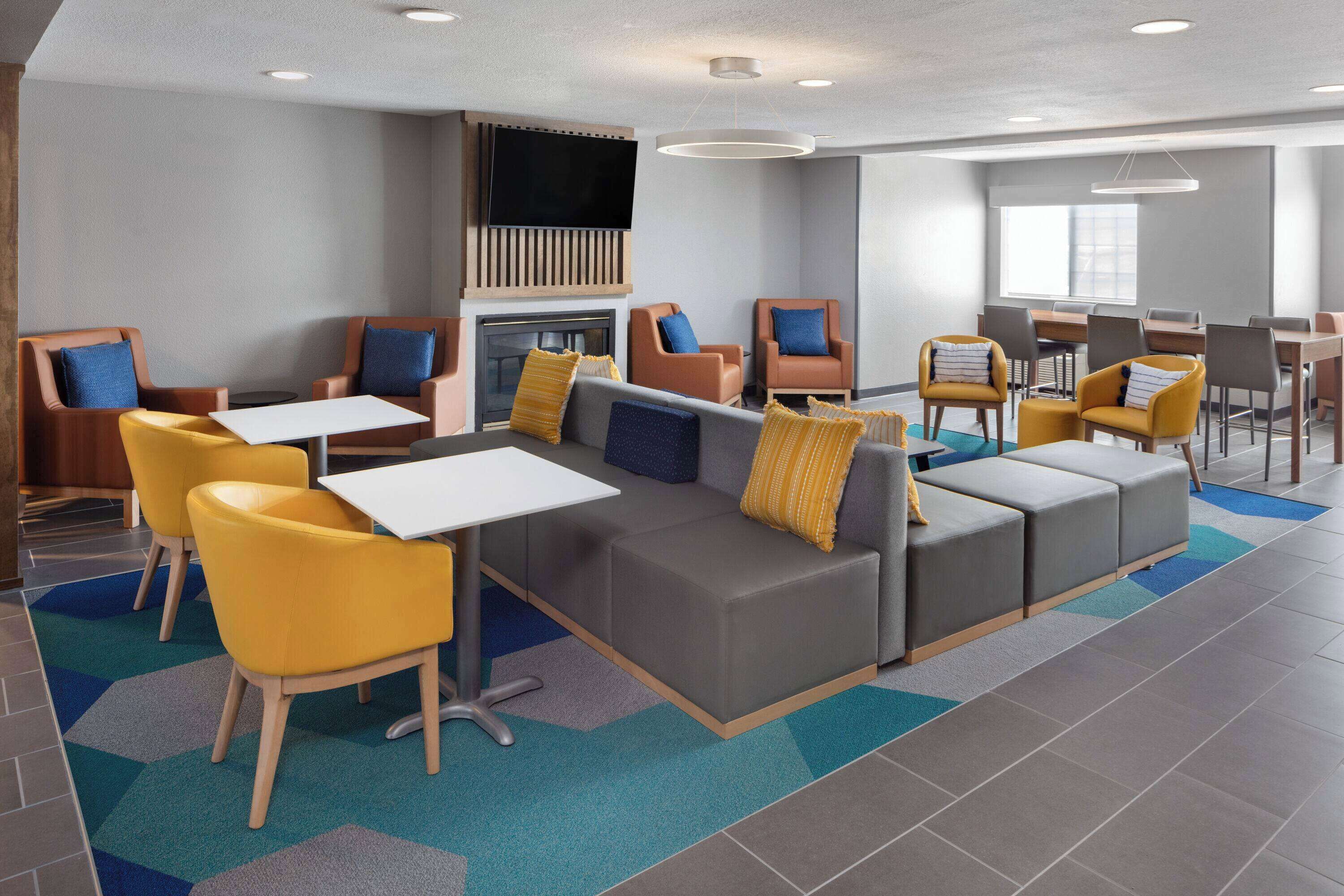 A modern hotel lobby with a gray sofa, yellow chairs, a flat-screen TV, and a fireplace.