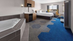 Una habitación de hotel moderna con una bañera grande, un dormitorio independiente con cama y escritorio, una pequeña cocina y vistas a la ciudad.
