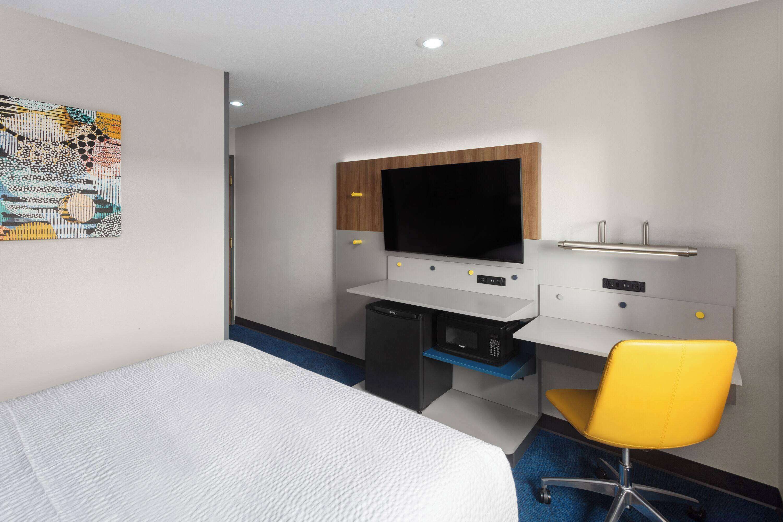 A hotel room with a bed, a desk with a flat-screen TV, a microwave, and a yellow chair.