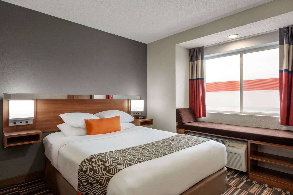 A hotel room with a bed, bedside tables, a bench, a TV, and a window with curtains.