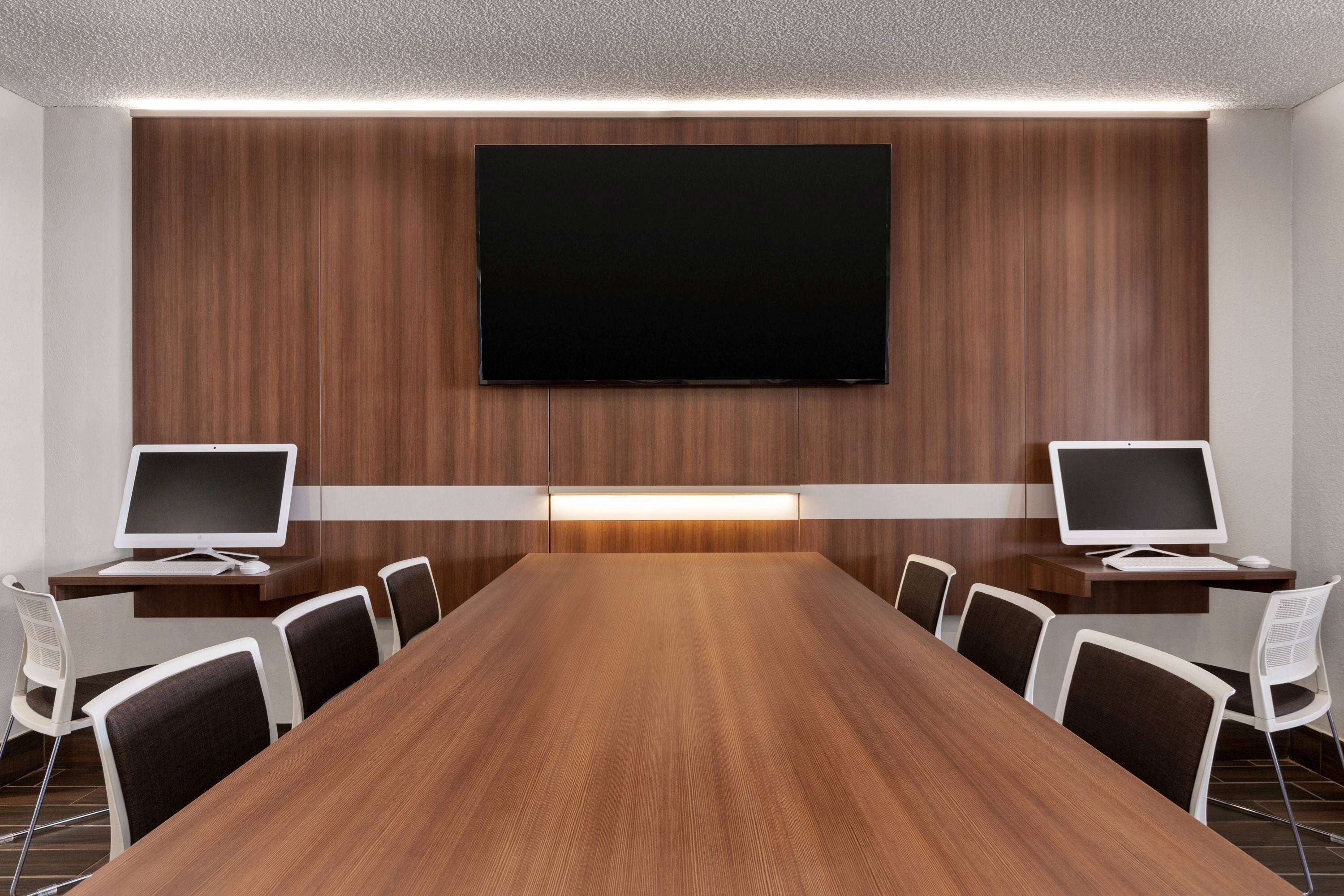 A conference room with a long wooden table, two monitors, and white chairs.
