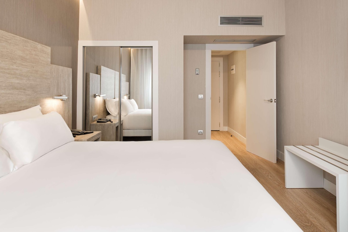Una habitación de hotel moderna con una cama grande, una mesita de noche, un televisor montado en la pared y un vestidor.