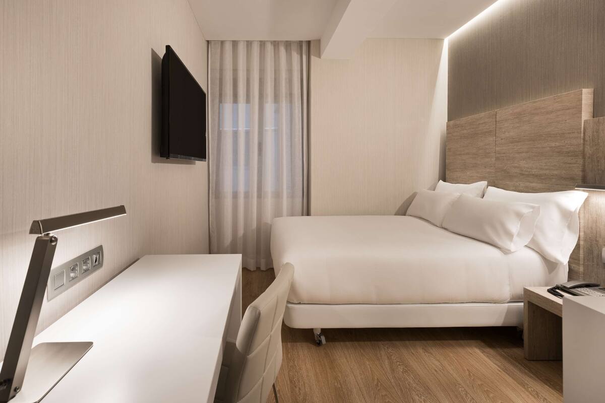 Moderni hotellihuone, jossa on sänky, yöpöytä, lamppu ja seinälle asennettu taulutelevisio.