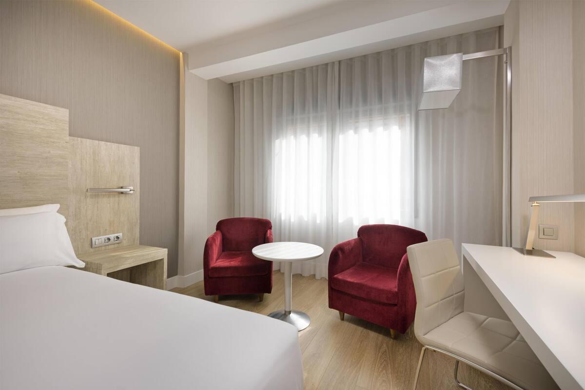 Hotellihuone, jossa on sänky, kaksi punaista nojatuolia, pieni pyöreä pöytä, työpöytä ja suuri ikkuna, jossa on ohuet verhot.