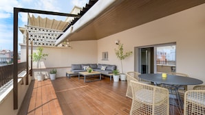 A balcony with a wooden floor, a sofa, a table, wicker chairs, and potted plants.