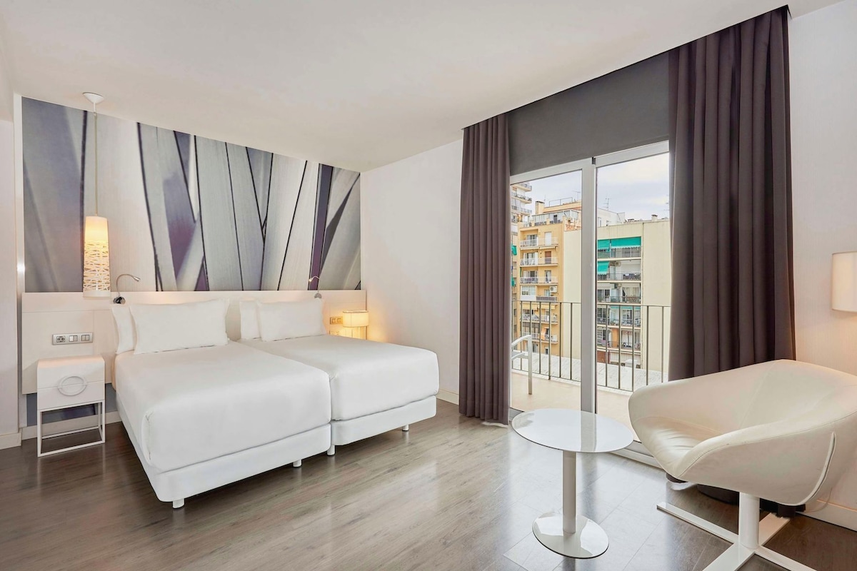 A hotel room with a bed, bedside tables, a chair, a balcony with a view of buildings, and a modern abstract painting on the wall.