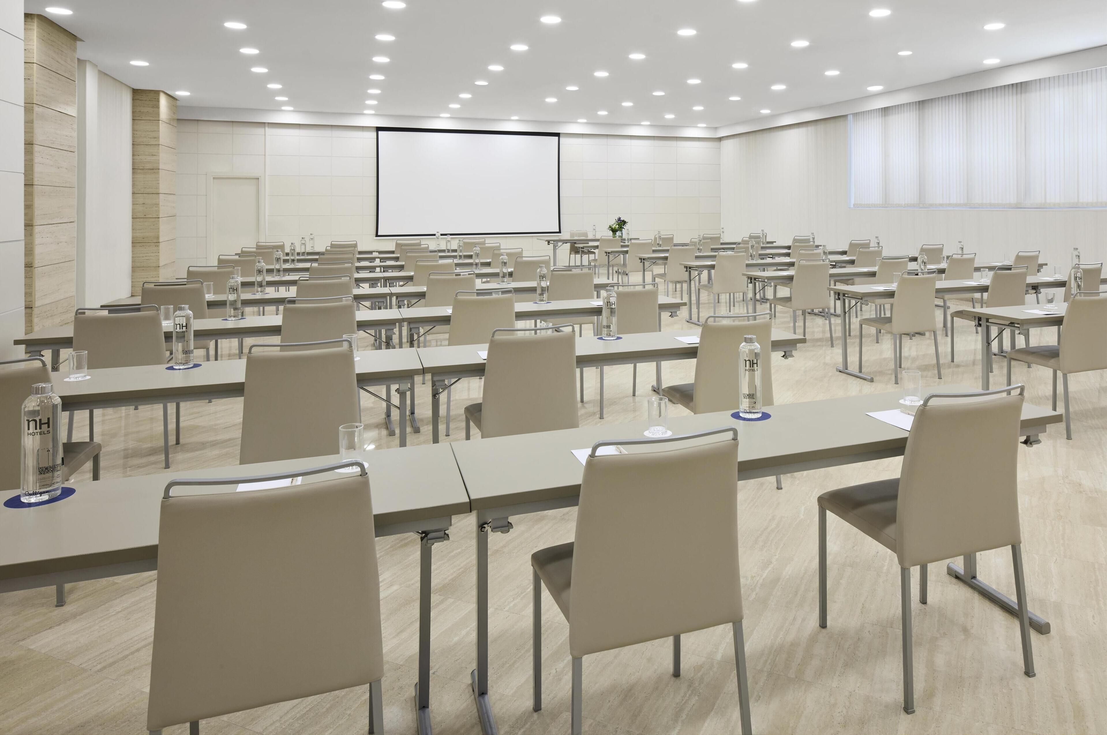 A conference room with multiple tables and chairs, a projector screen, and a large window with blinds.