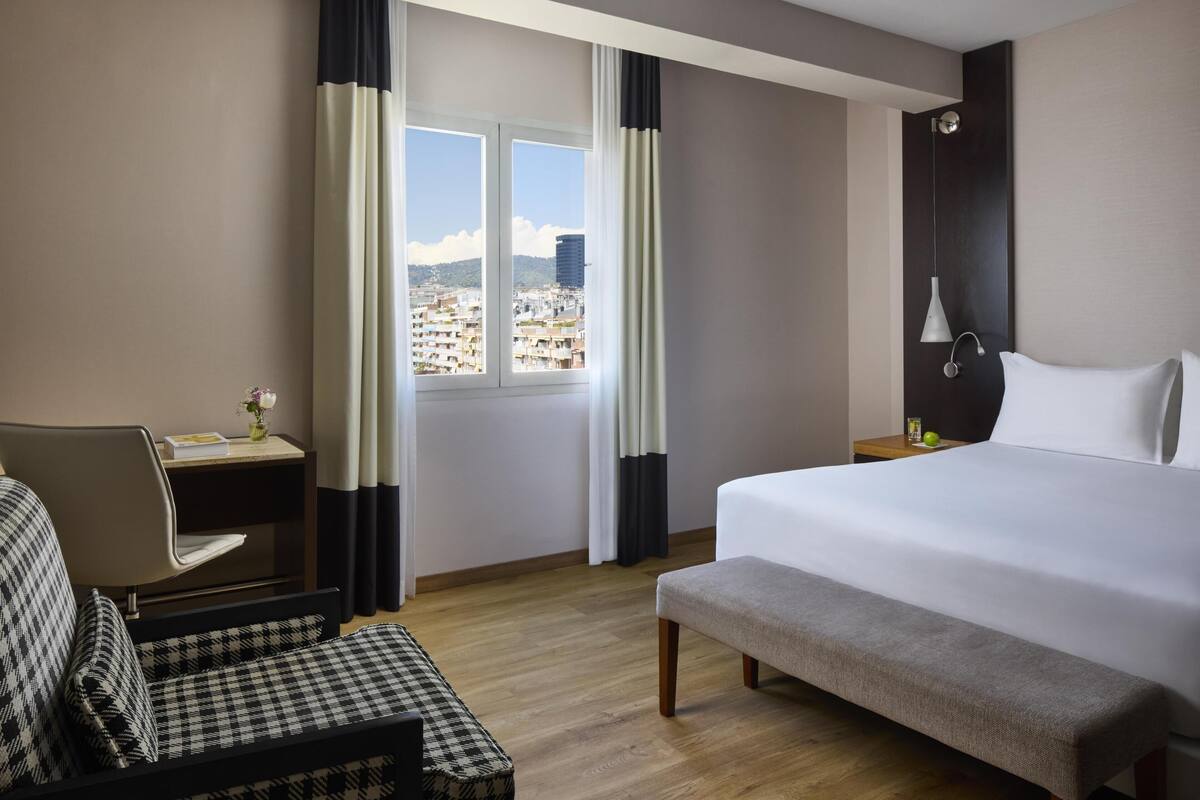 A hotel room with a bed, a desk, a chair, and a view of the cityscape through the window.