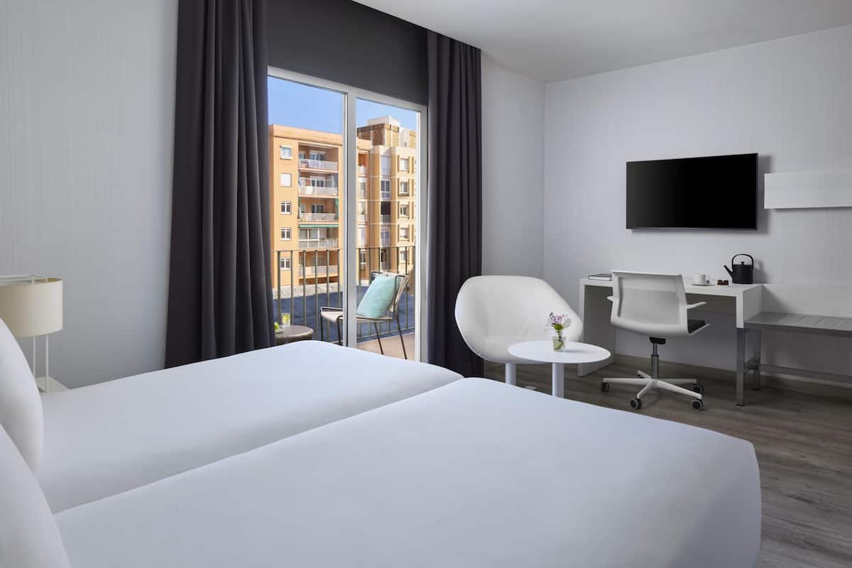 A hotel room with two beds, a desk with a chair, a TV, and a view of buildings through large windows.