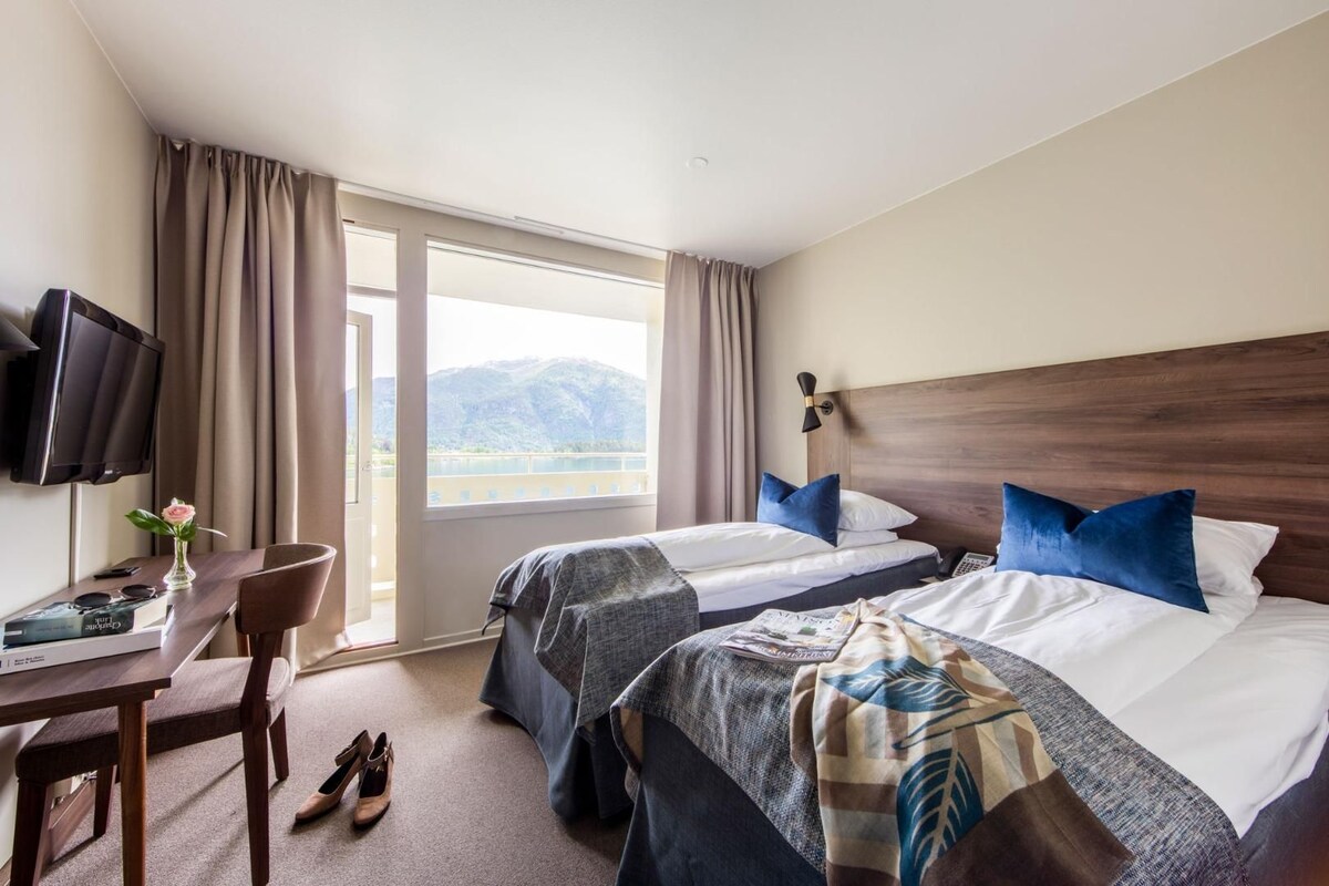 A hotel room with two beds, a desk with a chair, a TV, and a view of mountains.
