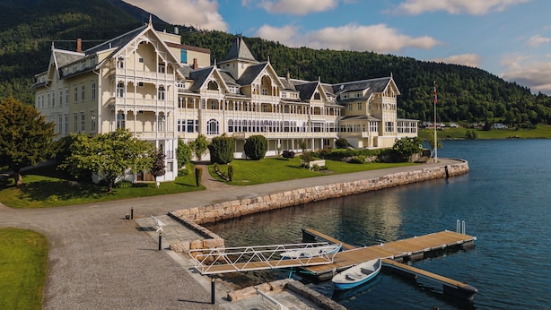 A large, multi-story building with a waterfront dock, surrounded by greenery and mountains.