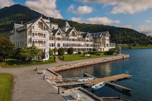 Ein großes, mehrstöckiges Gebäude mit einer Anlegestelle am Wasser, umgeben von Grünflächen und Bergen.