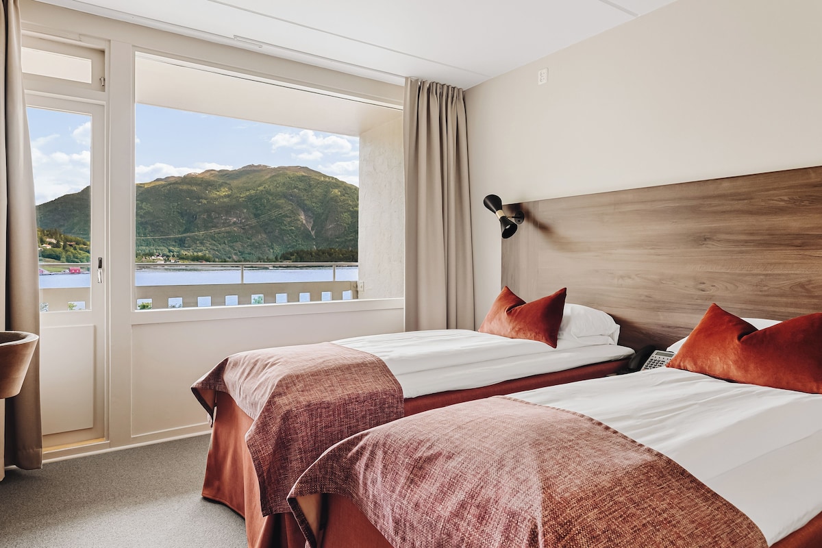 A hotel room with two beds, a wooden headboard, a view of a lake and mountains, and a curtain.