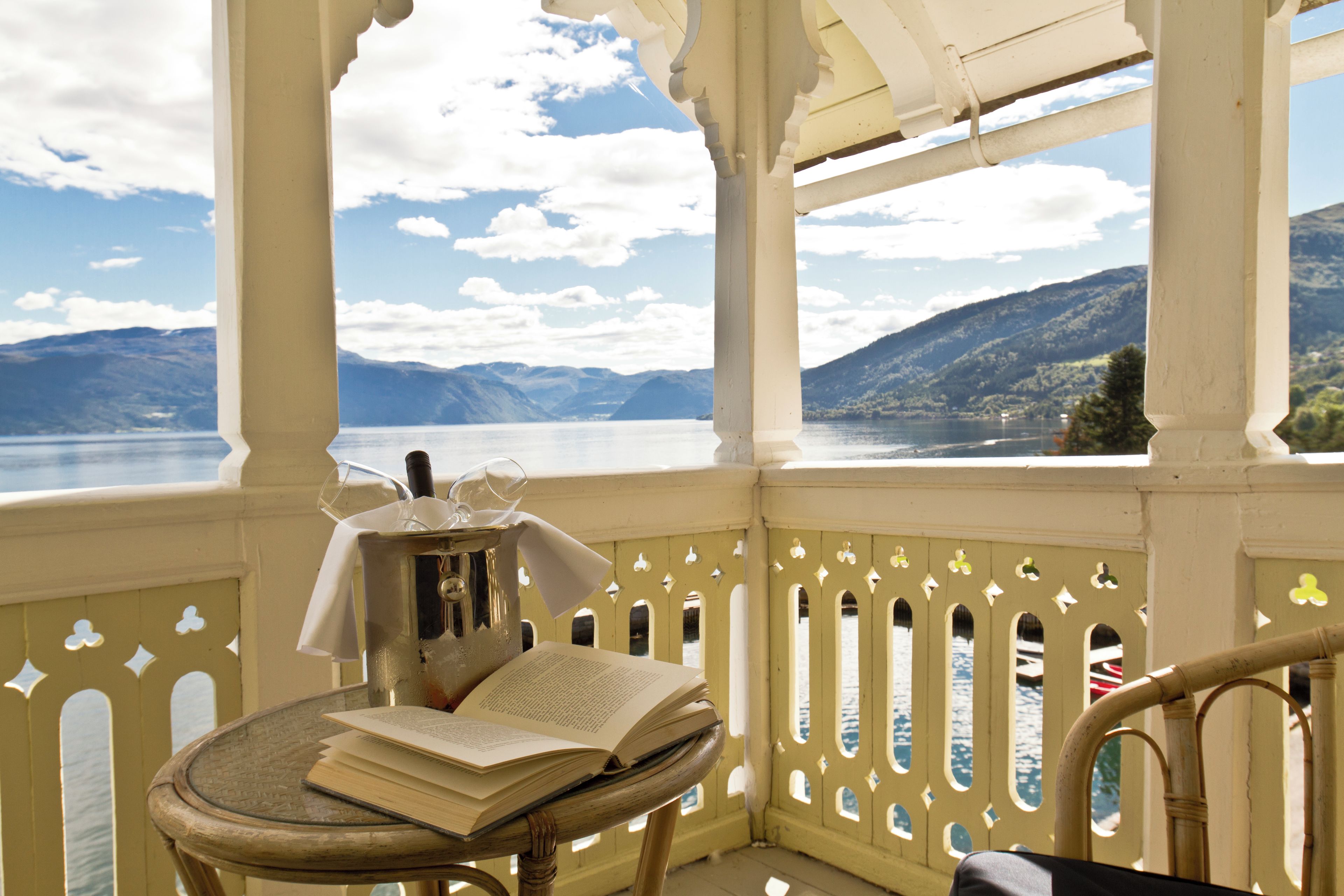 Ein Balkon mit Tisch, auf dem ein Buch und ein Glas stehen, mit Blick auf einen See und Berge.
