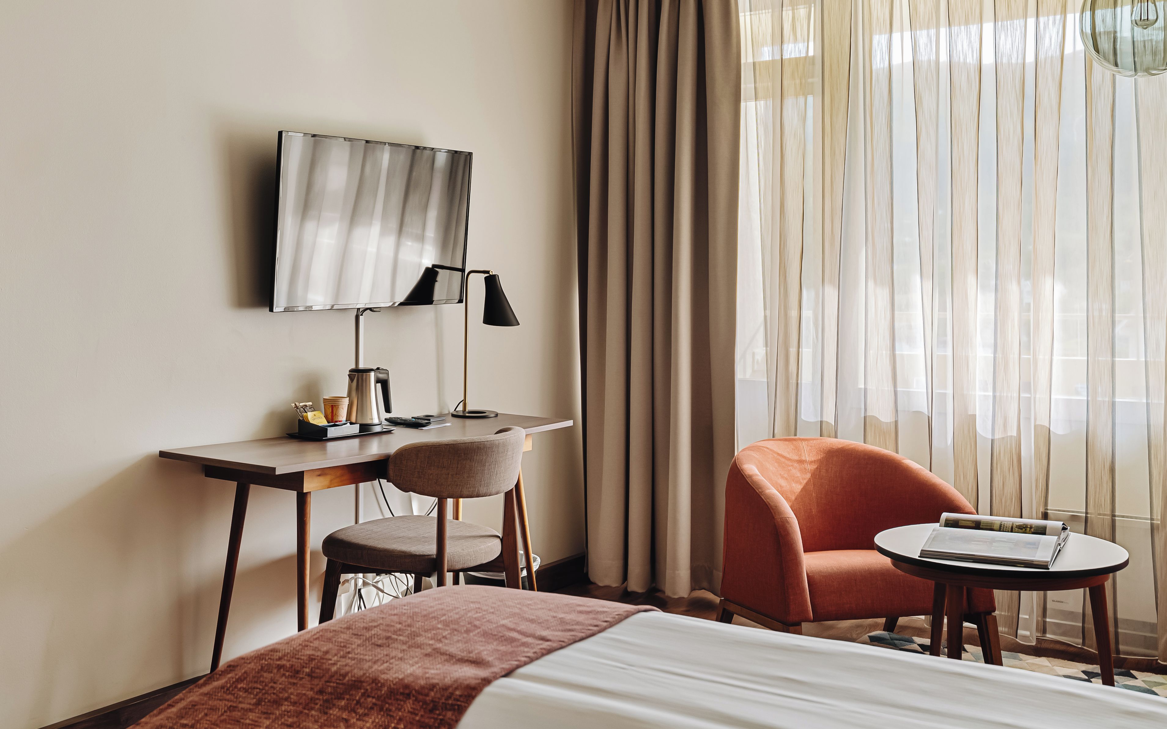 Una camera d'albergo con un letto, una scrivania con televisione a schermo piatto, una sedia, un tavolino con una rivista e tende trasparenti.