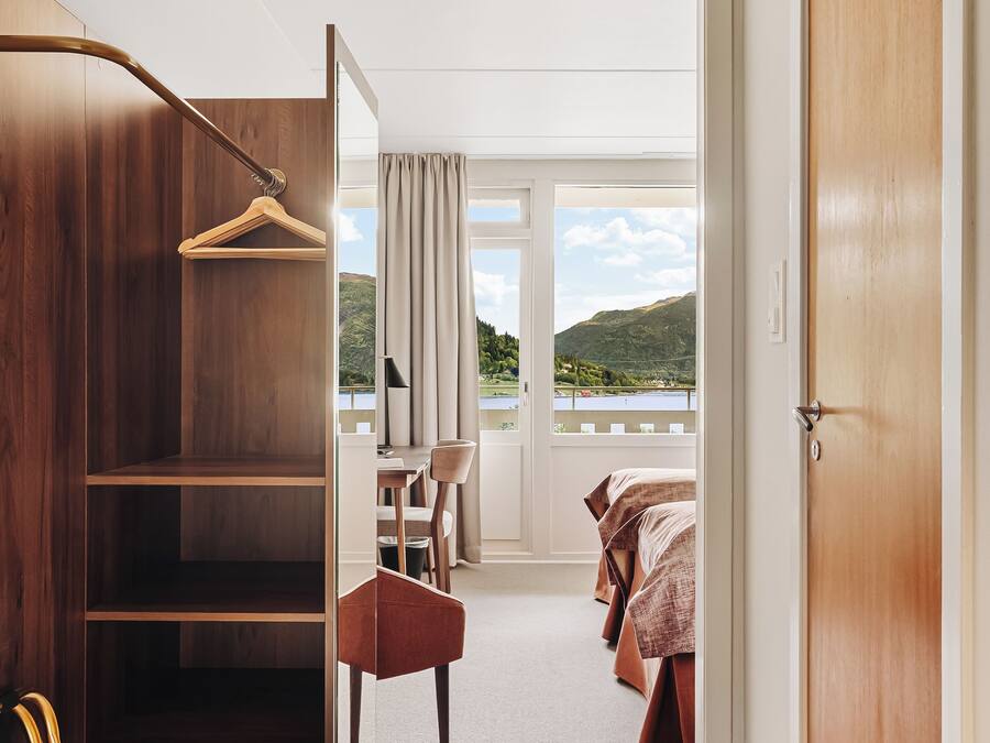 A hotel room with a wooden wardrobe, a desk with a chair, a bed, and a view of a lake.