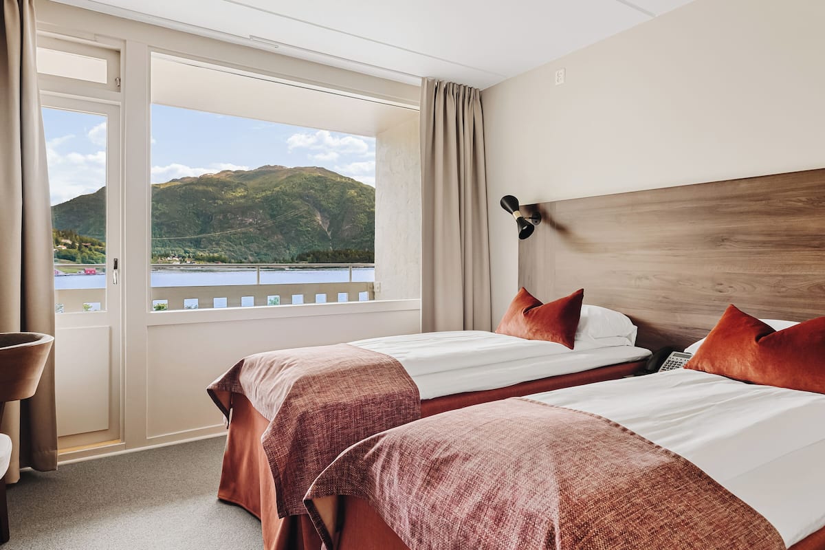 Habitación de hotel con dos camas, cabecero de madera, vista a un lago y montañas, y una cortina.