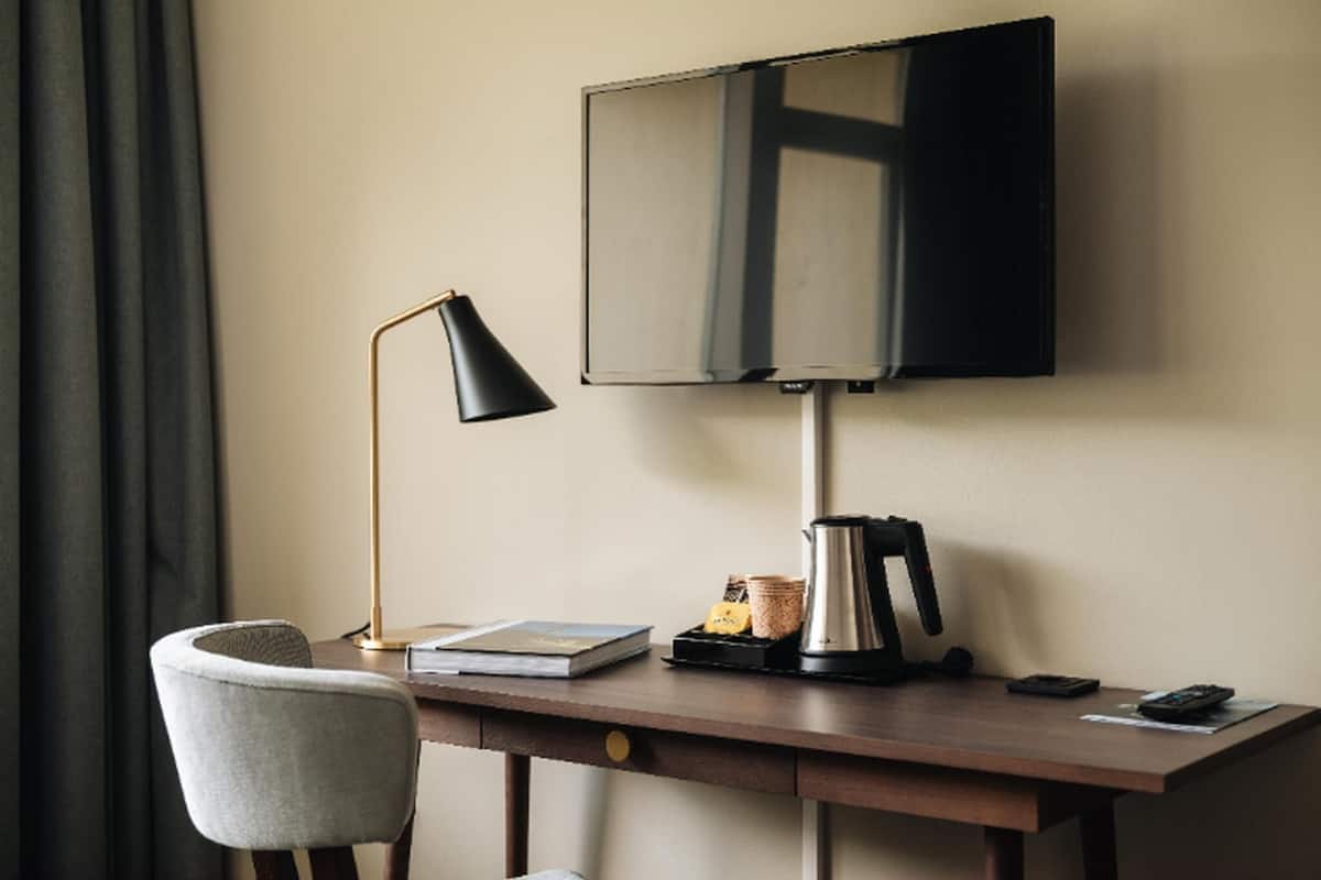 Habitación de hotel con escritorio, silla, televisor de pantalla plana montado en la pared, lámpara, cafetera y control remoto.