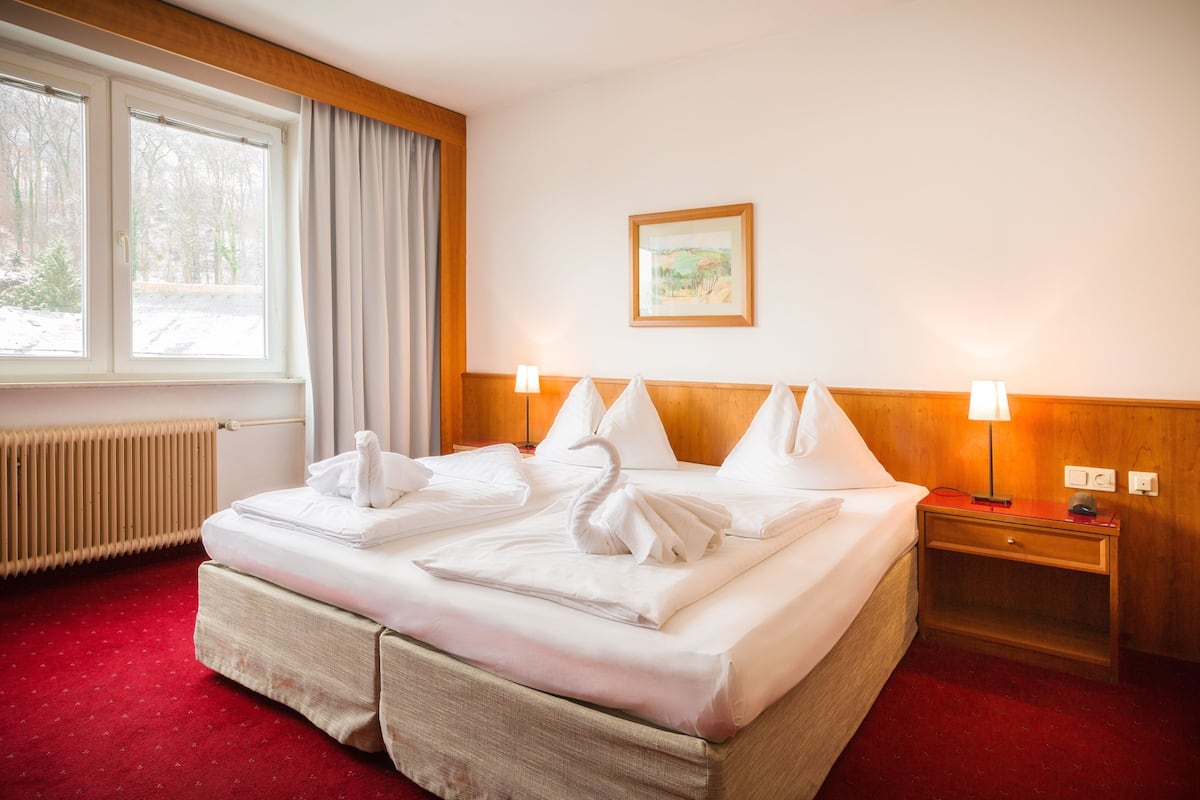 A hotel room with a neatly made bed, a wooden headboard, a bedside table with a lamp, a radiator, and a window with curtains.