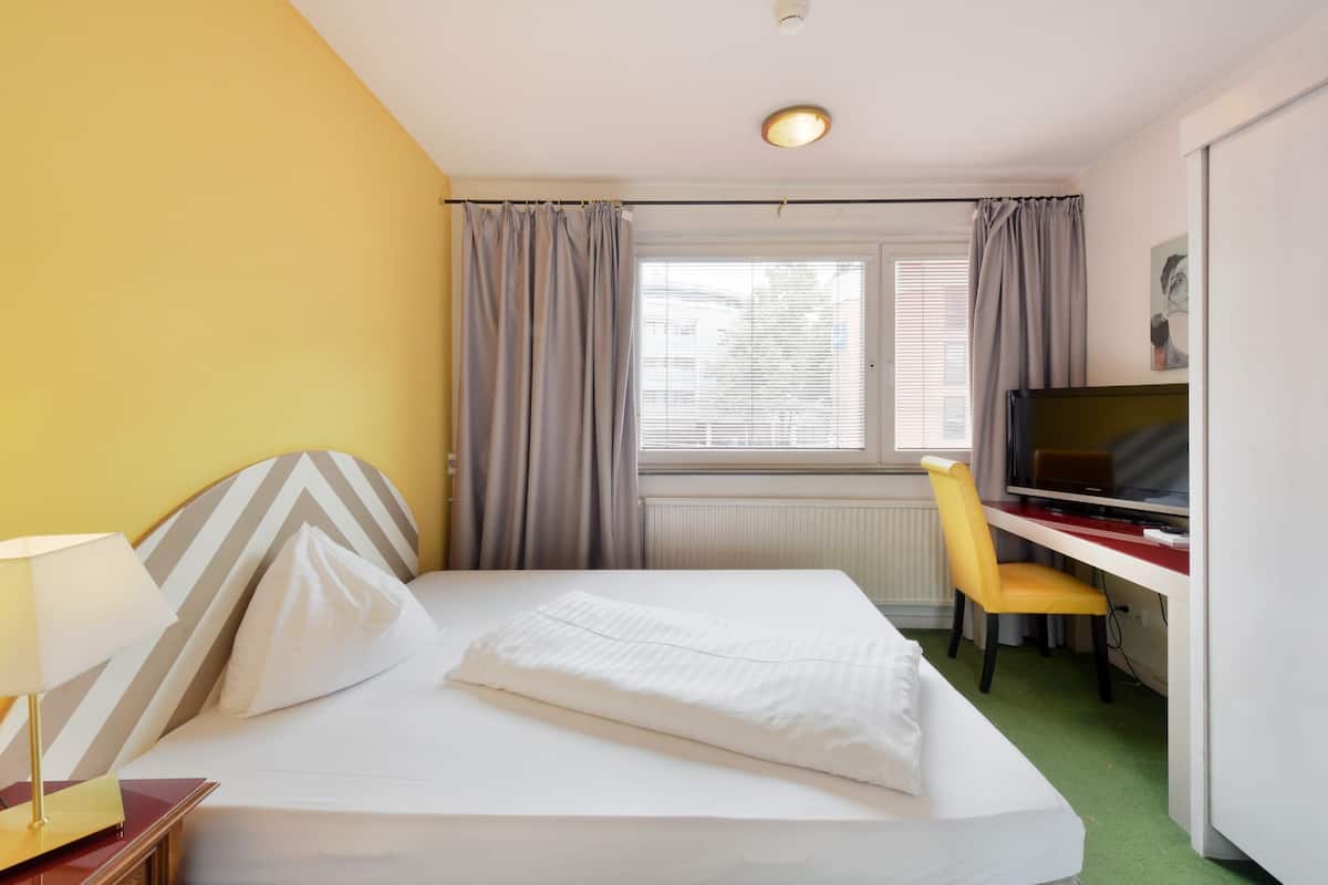 A hotel room with a bed, a desk with a chair, a TV, and a window with curtains.