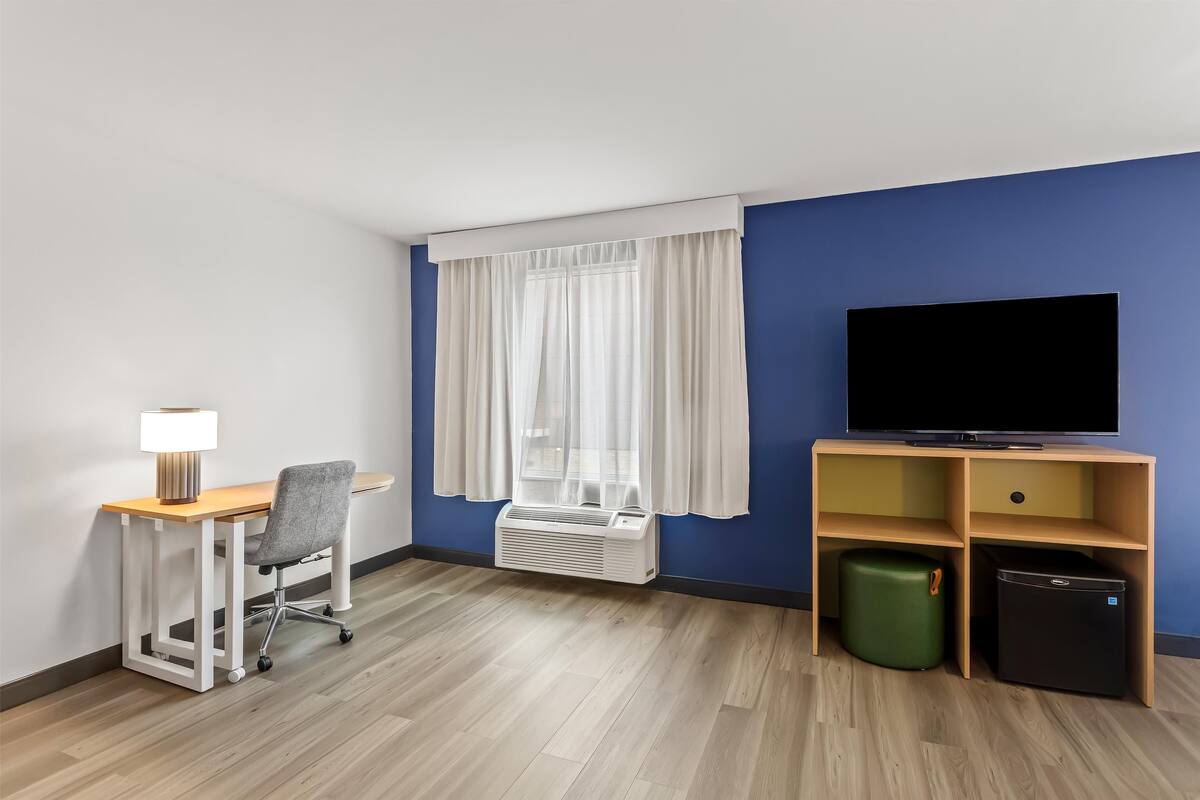 Habitación de hotel con escritorio de madera, una silla, televisor, calentador, cubo de basura y ventana con cortinas.