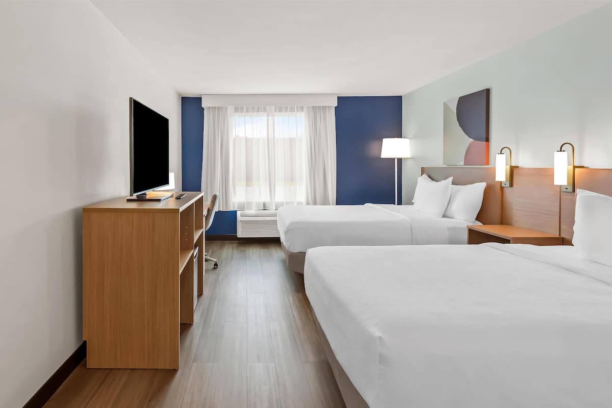 Una habitación de hotel con una cama, un escritorio con televisor, una lámpara y un ventanal con cortinas.