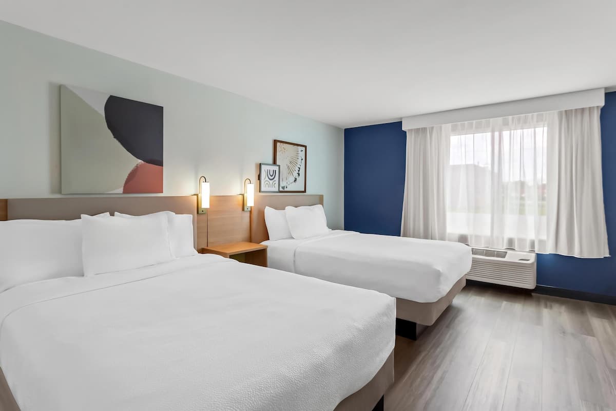 Habitación de hotel con dos camas, una mesita de noche, decoración en la pared, una ventana con cortinas y un equipo de aire acondicionado.