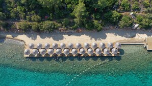 An aerial view of a beach with a wooden deck with umbrellas, clear turquoise water, and a forested area in the background. - Hapimag Resort Sea Garden (Bodrum)