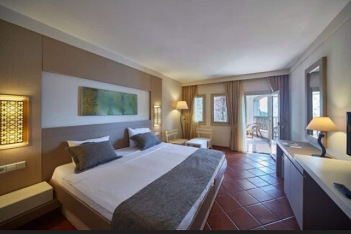 A hotel room with a large bed, bedside tables, a desk, and a balcony with curtains.