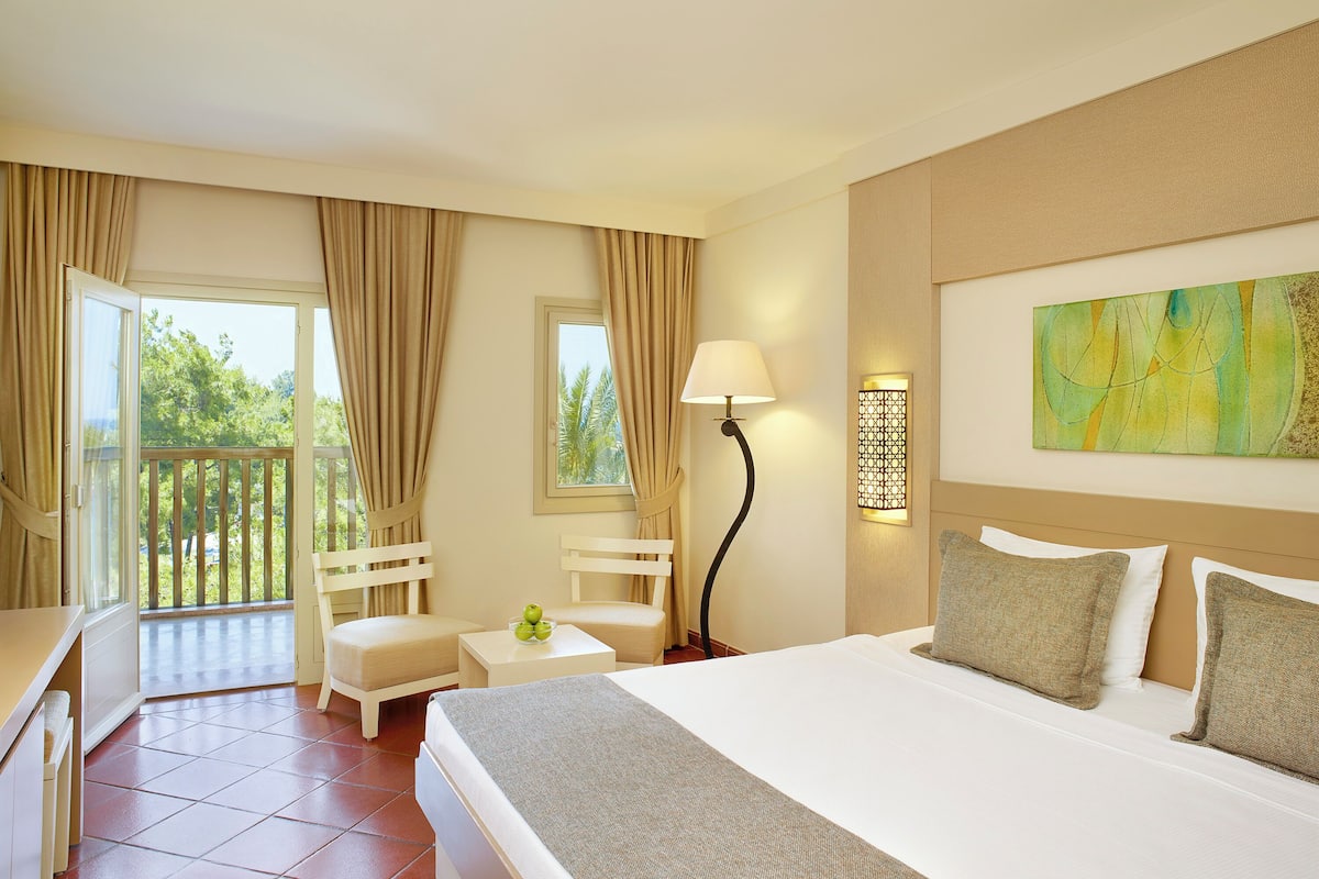 A hotel room with a bed, a desk, a chair, a small table with a lamp, and a balcony with curtains.