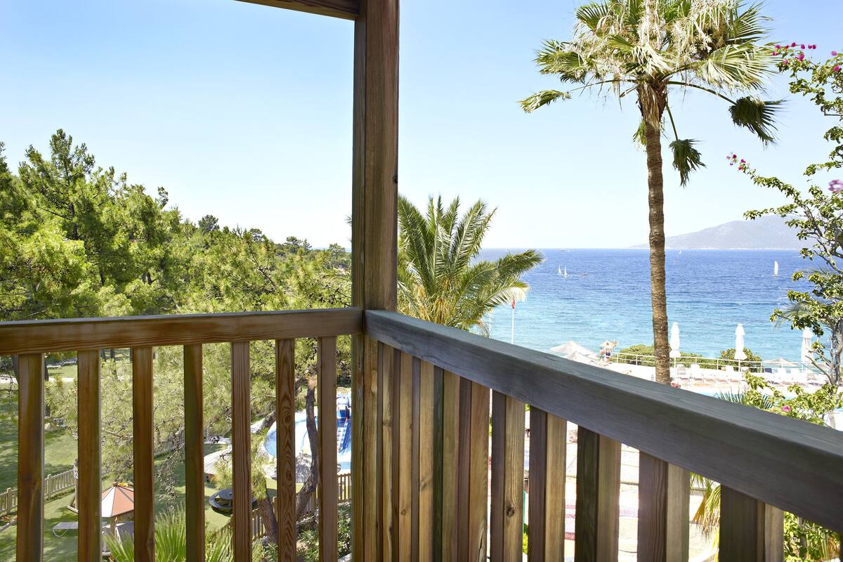 A wooden balcony overlooking a pool area with a view of the sea and boats.