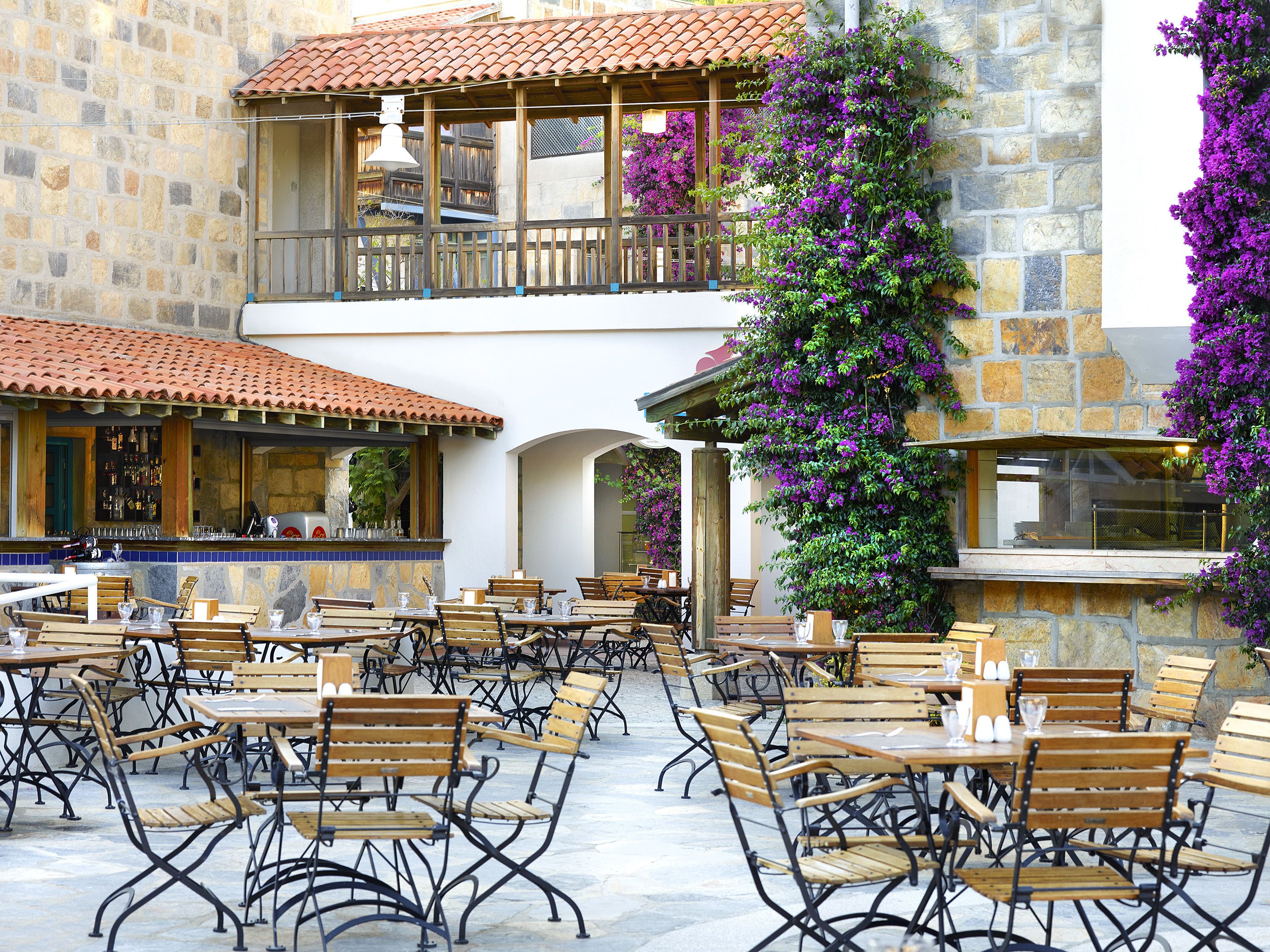 An outdoor café with multiple wooden tables and chairs, stone walls, and a bar area with a tiled roof.