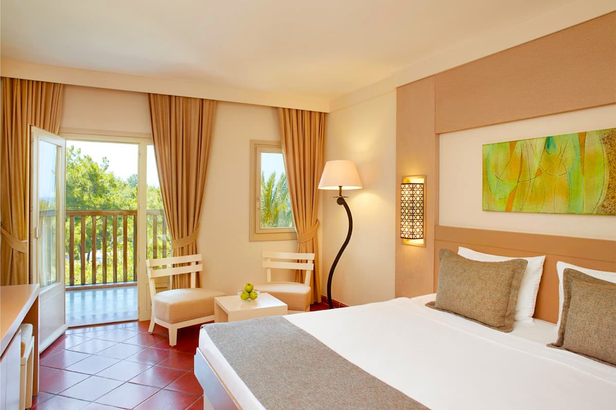 A hotel room with a bed, a desk, a chair, a small table, a lamp, and a balcony with curtains.