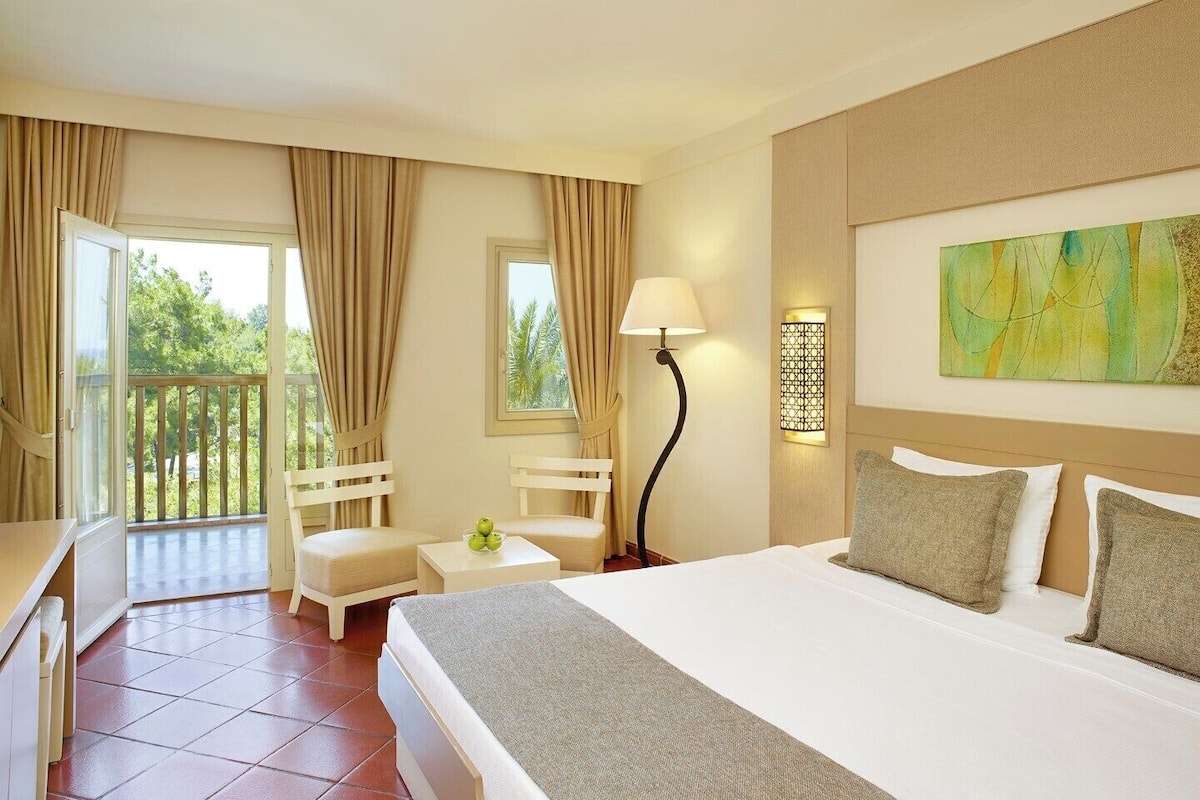 A hotel room with a bed, a desk, a chair, a lamp, and a balcony with curtains.