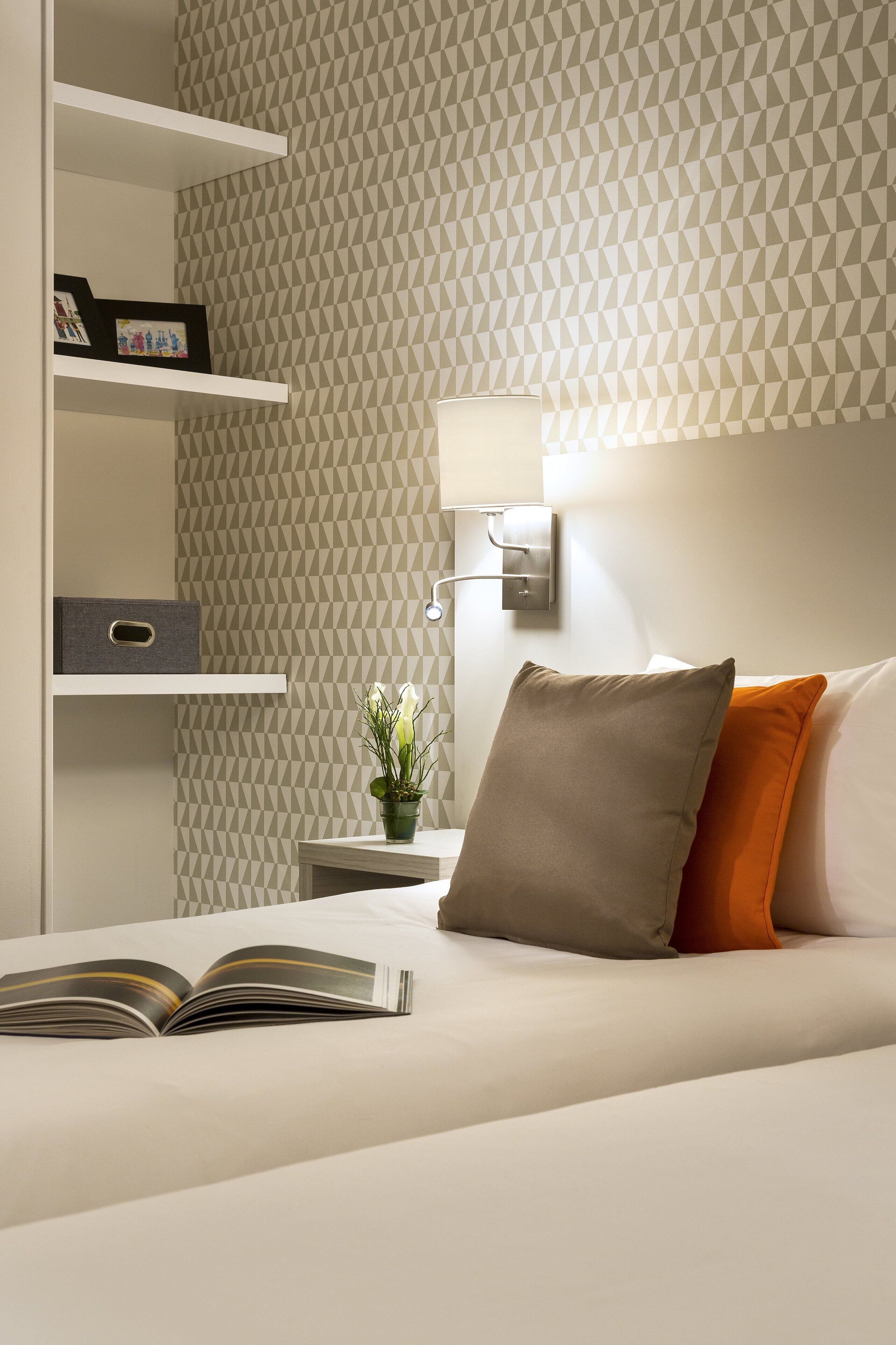 Une chambre d’hôtel bien rangée, avec un lit, une table de chevet dotée d’une lampe, une petite plante en pot et des étagères contenant des boîtes de rangement et des photos encadrées.