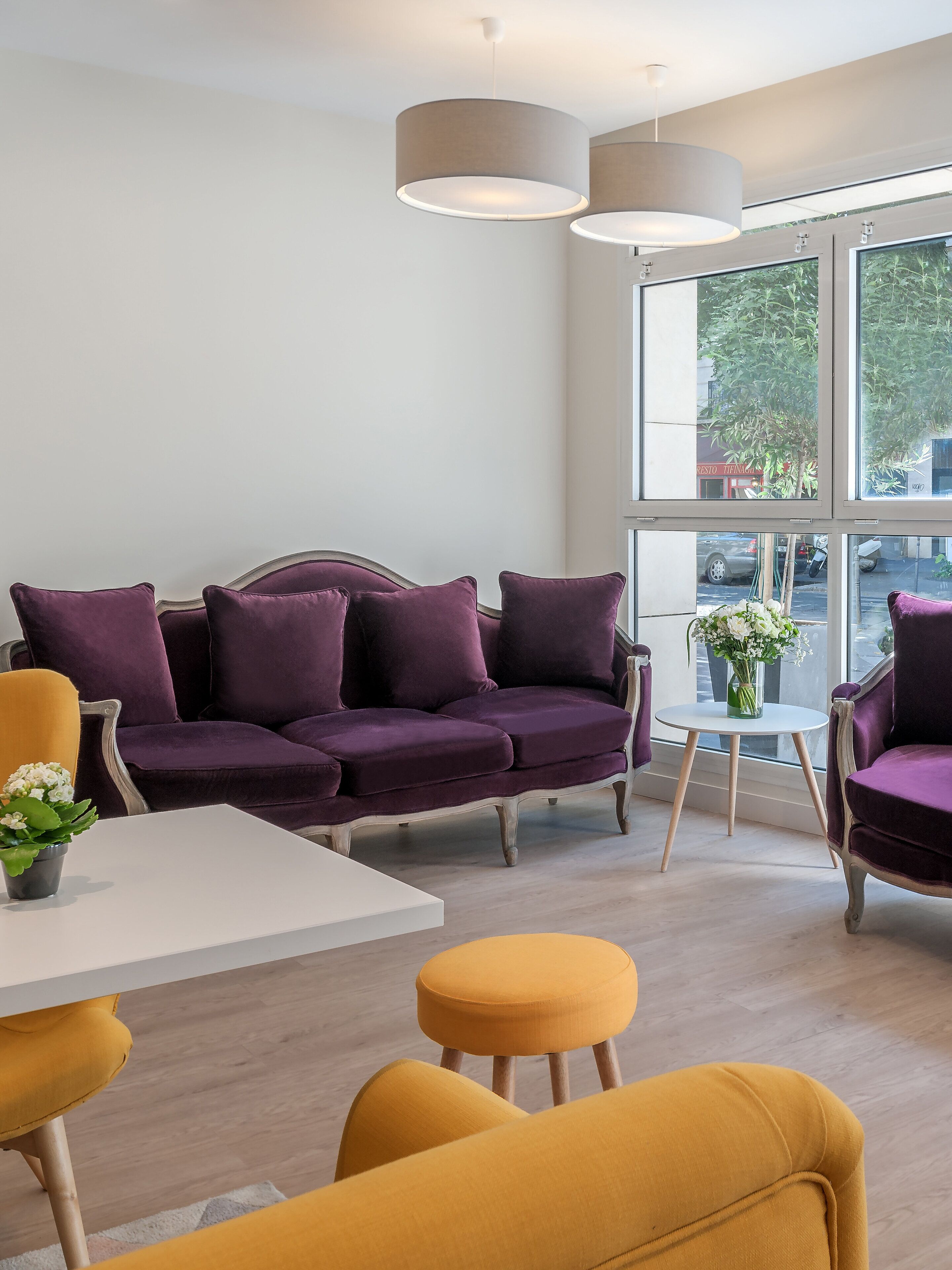 Una sala de estar moderna con un sofá púrpura, sillas amarillas, una mesa blanca y ventanas amplias con vistas a la calle.